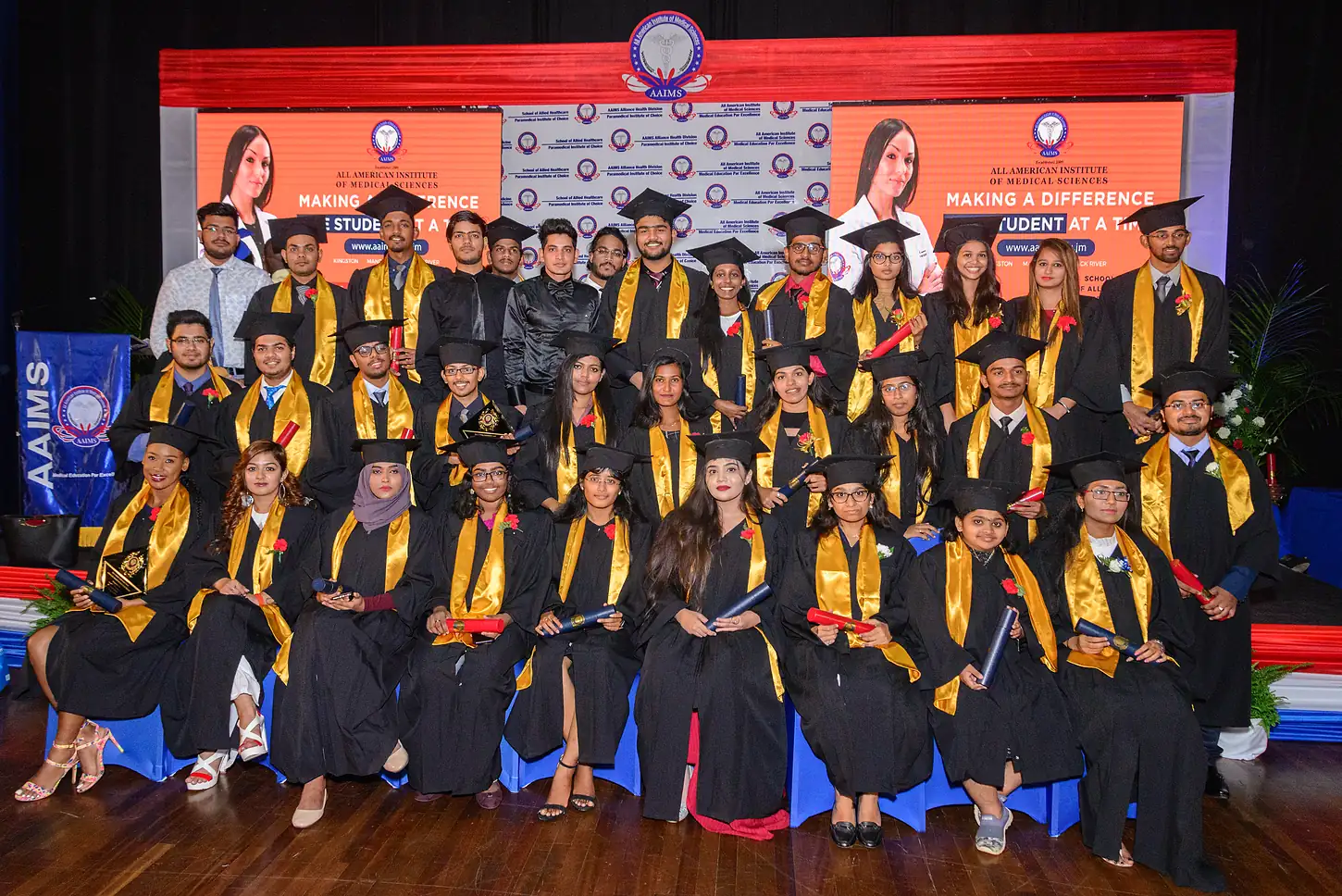 A graduating class of AAIMS students in black gowns and gold stoles on stage beneath the AAIMS crest and a Making a Difference banner.